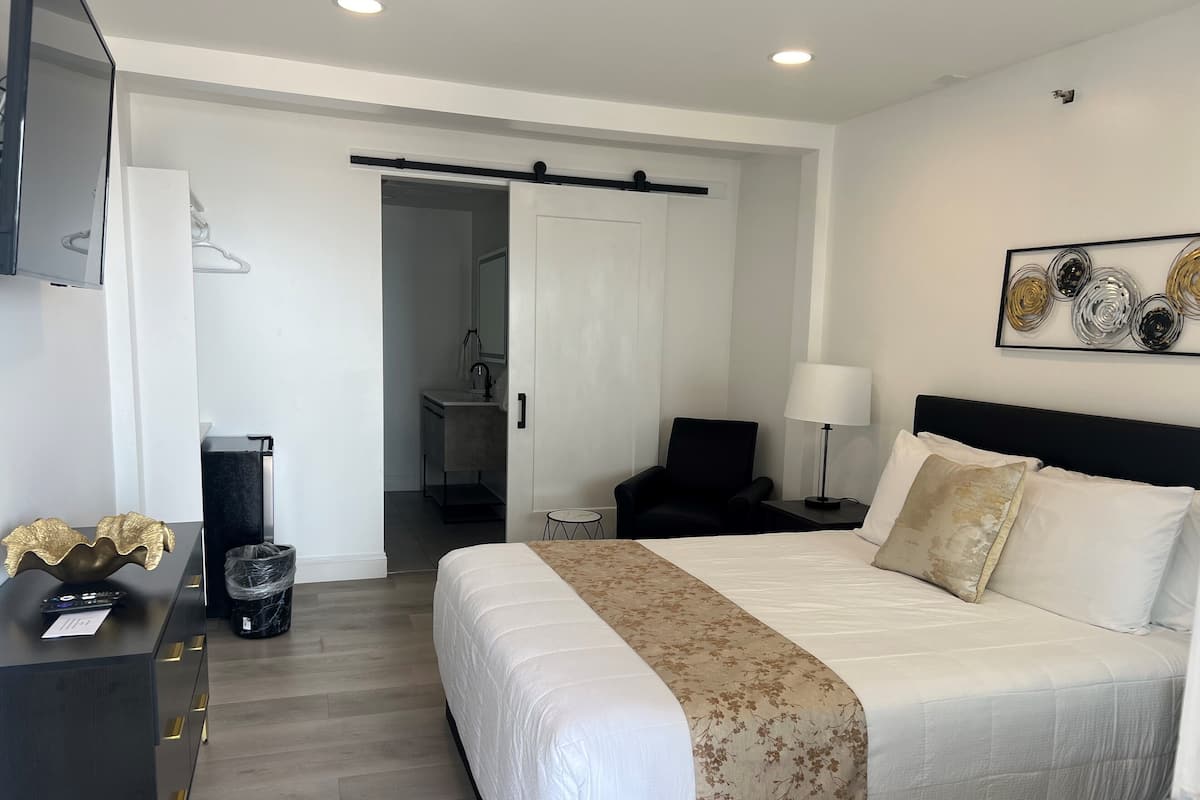 A modern hotel room with a large bed, a desk with a lamp, a chair, and a bathroom visible through an open door.