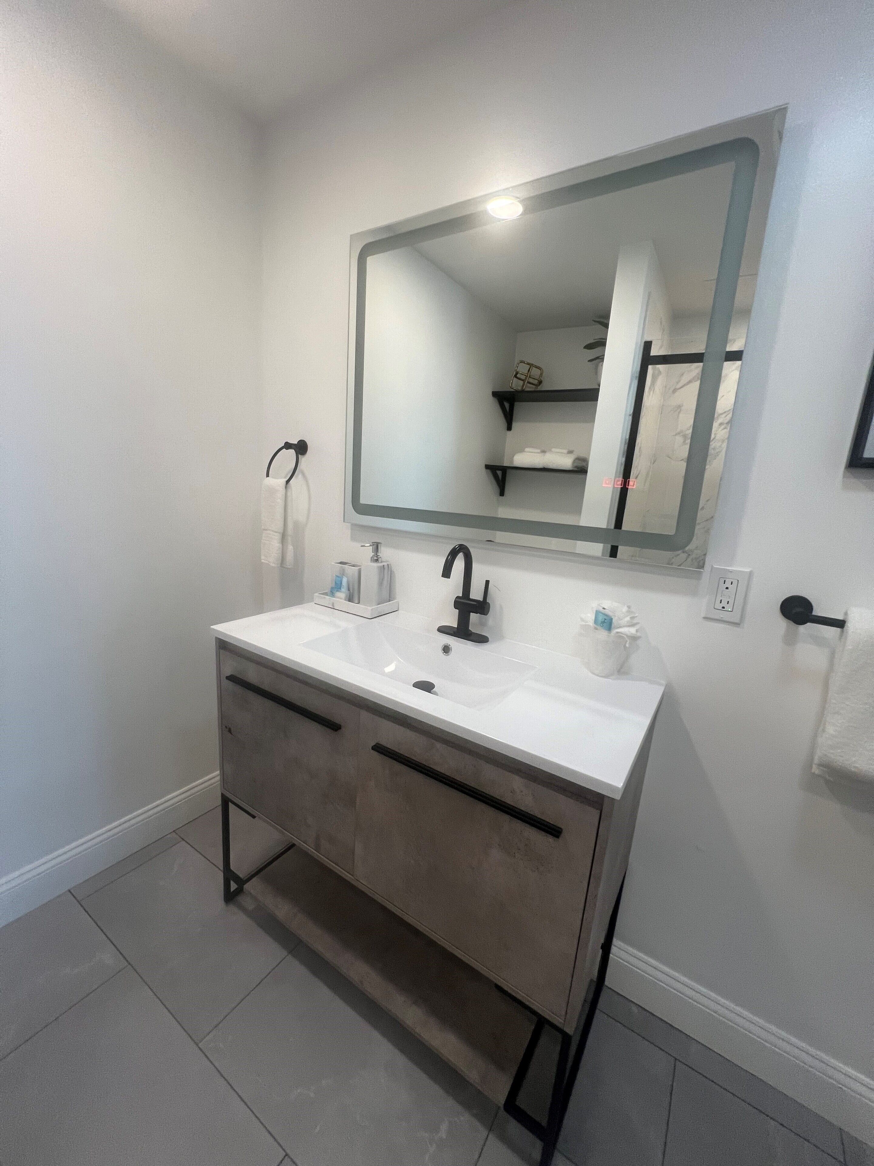 Un baño moderno con un lavamanos blanco, grifo negro y un espejo grande. Hay dos toalleros y un estante con toallas.