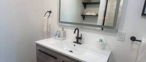 Un baño moderno con un lavamanos blanco, grifo negro y un espejo grande. Hay dos toalleros y un estante con toallas.