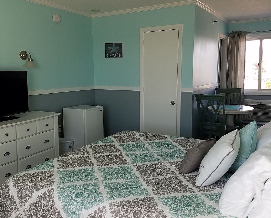 Un dormitorio con cama, cómoda, televisor, refrigerador pequeño y ventana con cortinas.