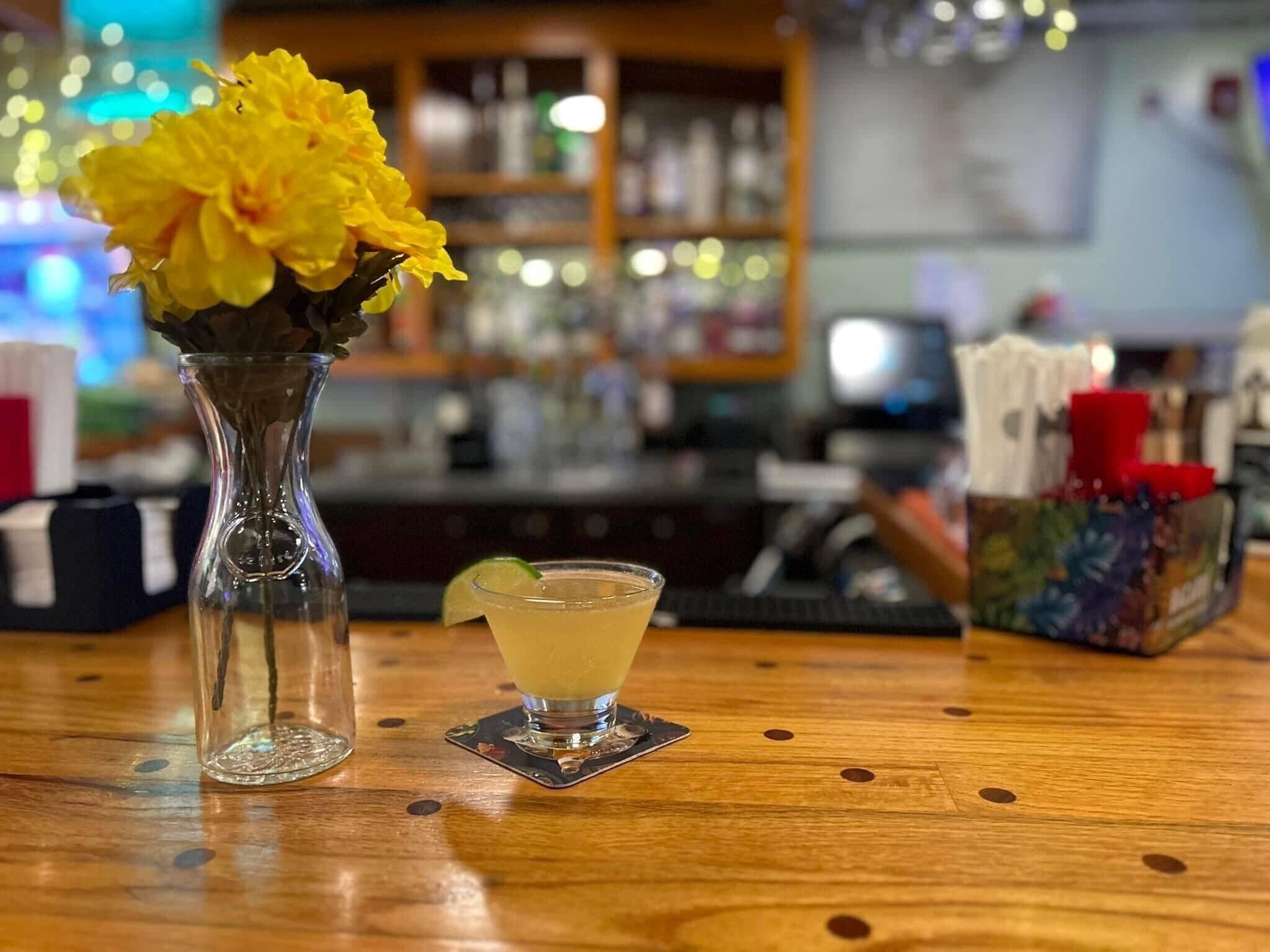 Un bar con un jarrón de vidrio transparente lleno de flores amarillas, una copa de cóctel con una rodaja de lima y un posavasos sobre la barra de madera.