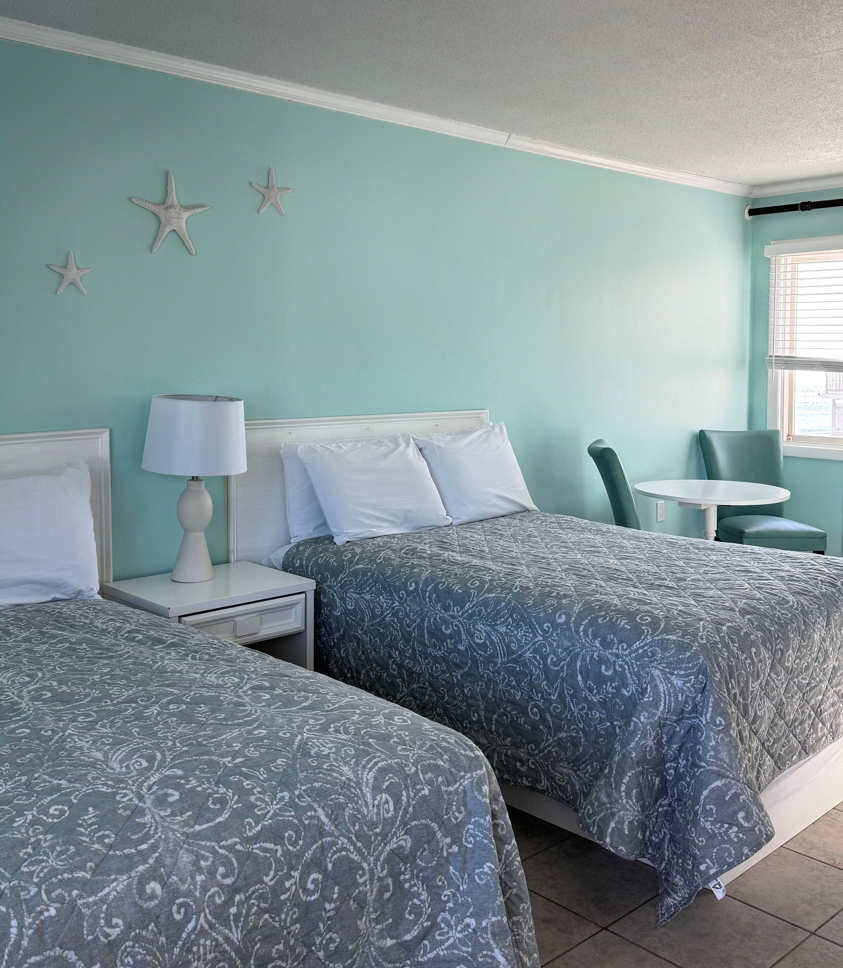 A room with two beds, a nightstand with a lamp, a small table, and a chair.