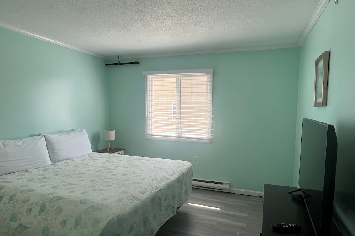A bedroom with a bed, a nightstand, a lamp, a window with blinds, a heater, and a TV.