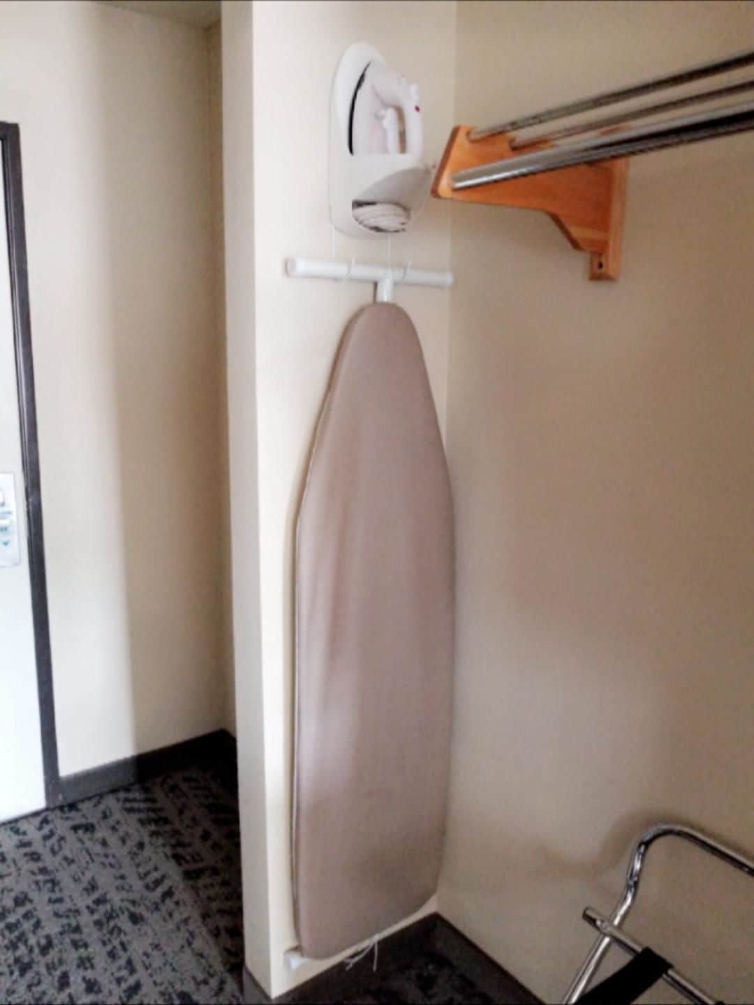 A corner of a hotel room with a hanging iron and ironing board, a wooden shelf with a mirror, and a patterned carpet.