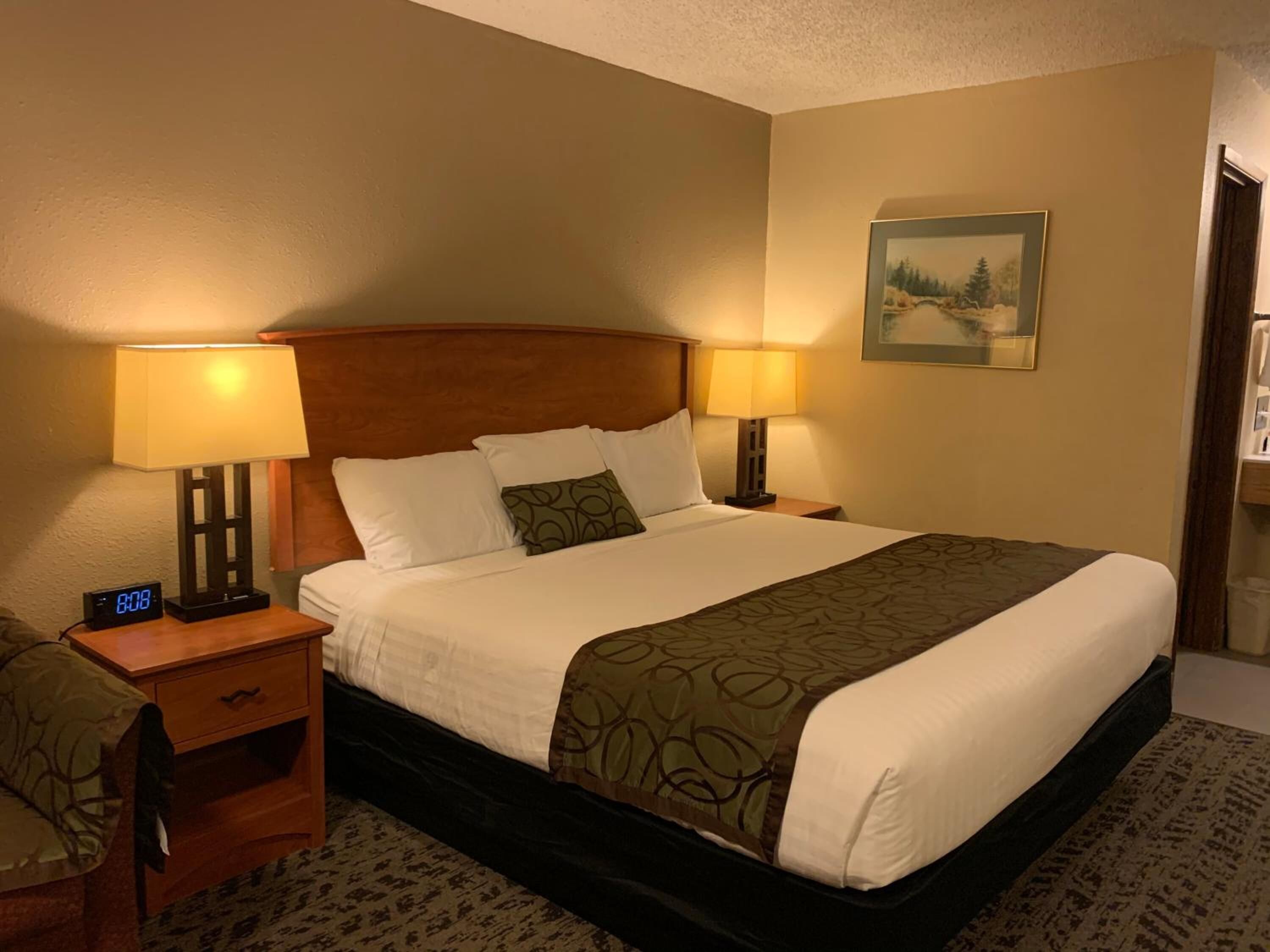 A hotel room with a large bed, two bedside tables with lamps, a nightstand, a clock, and a framed picture on the wall.