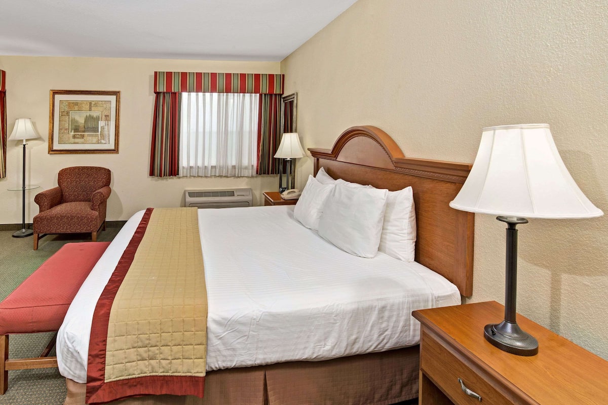 Una habitación de hotel con una cama, una silla, una mesita de noche con lámpara, un cuadro enmarcado en la pared y una ventana con cortinas.
