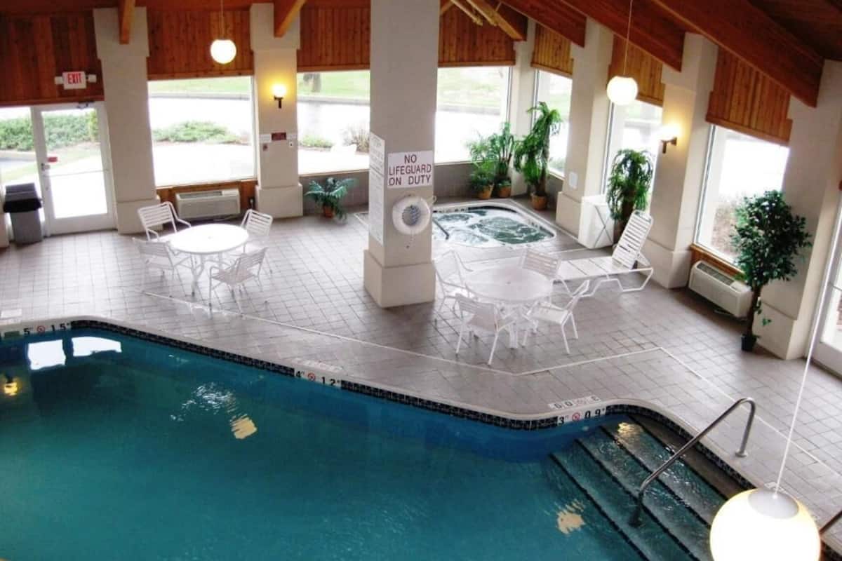 Una piscina interior con un jacuzzi, rodeada de sillas y mesas blancas, y un letrero que indica que no hay socorrista de servicio.