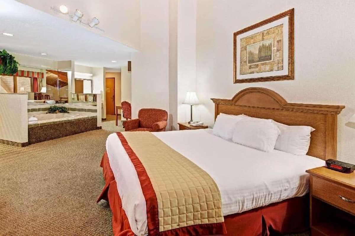 Habitación de hotel con una cama grande, cabecera de madera, mesita de noche, silla, espejo y un cuadro enmarcado en la pared.
