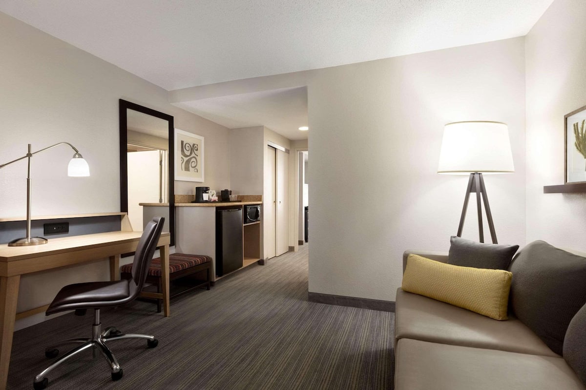 A hotel room with a desk, chair, sofa, lamp, and a small kitchenette.