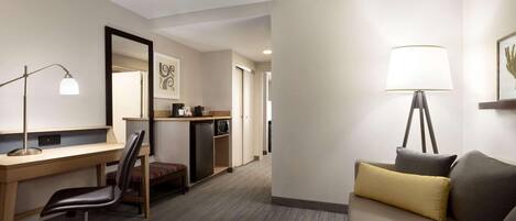 A hotel room with a desk, chair, sofa, lamp, and a small kitchenette.