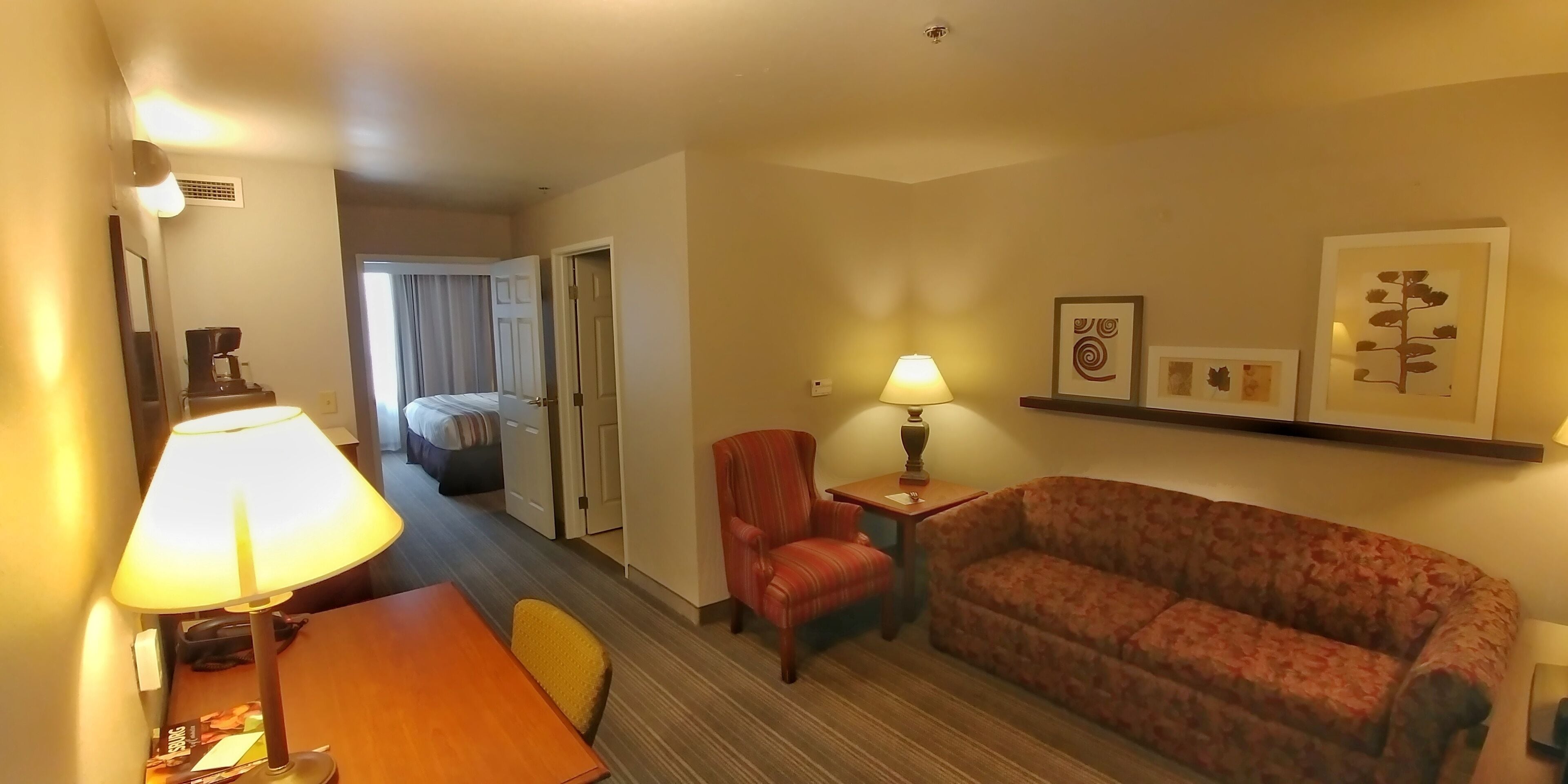 A hotel room with a wooden desk, a red armchair, a patterned sofa, a lamp, and a bed with curtains.