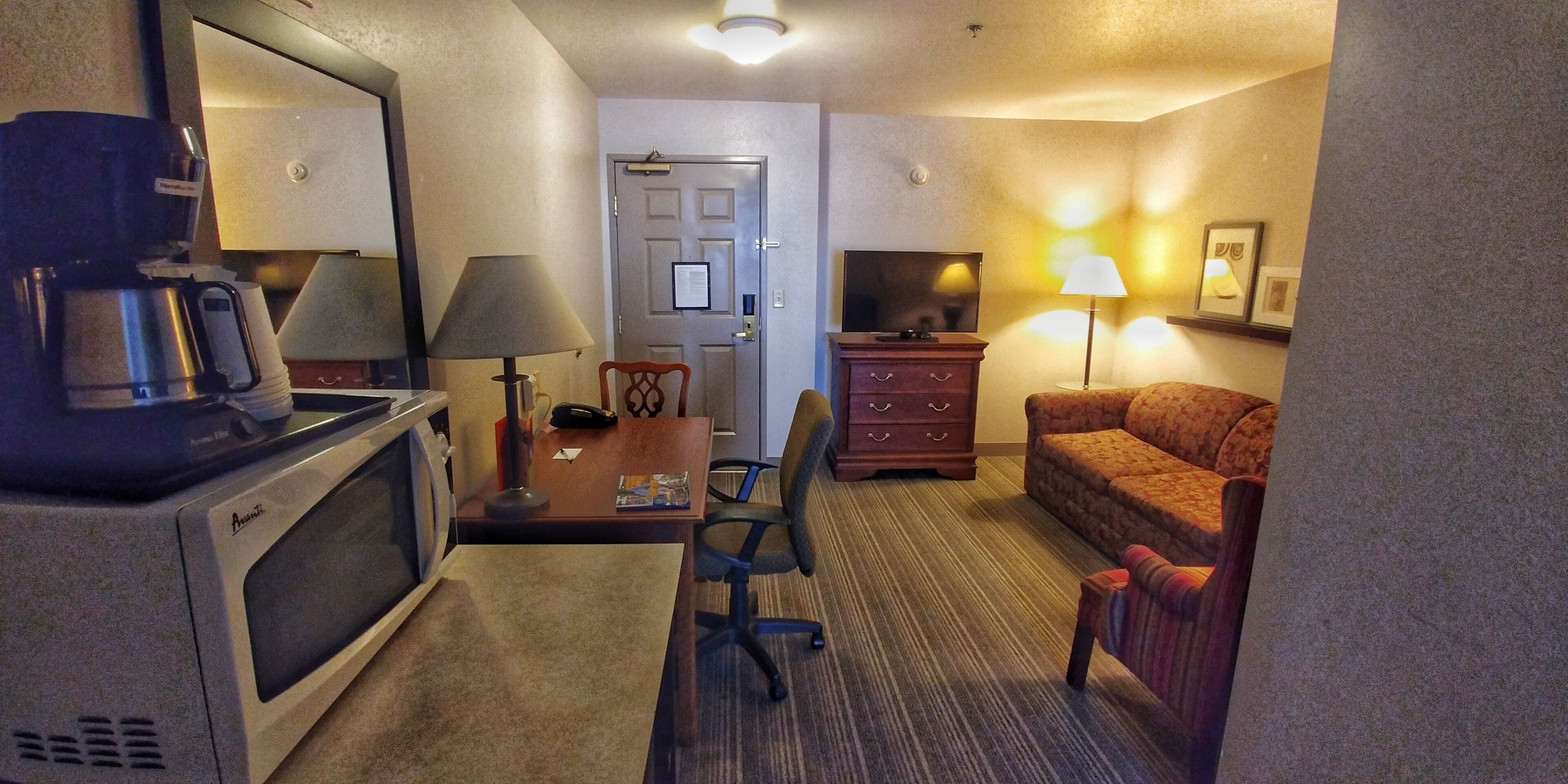 A hotel room with a kitchenette, a desk with a lamp, a chair, a sofa, and a television.