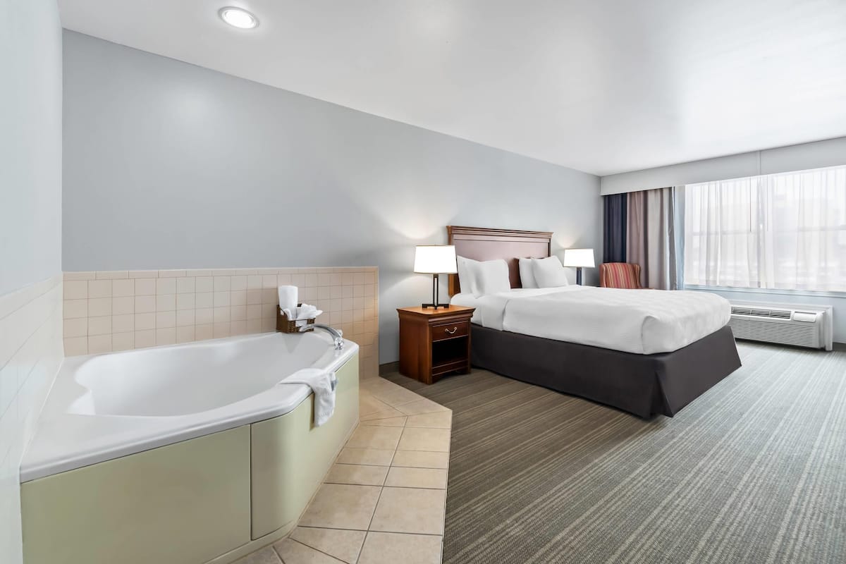 A hotel room with a bathtub, a double bed, a nightstand with a lamp, and a window with curtains.