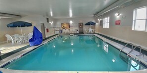 An indoor swimming pool with clear water, surrounded by white chairs and blue umbrellas.
