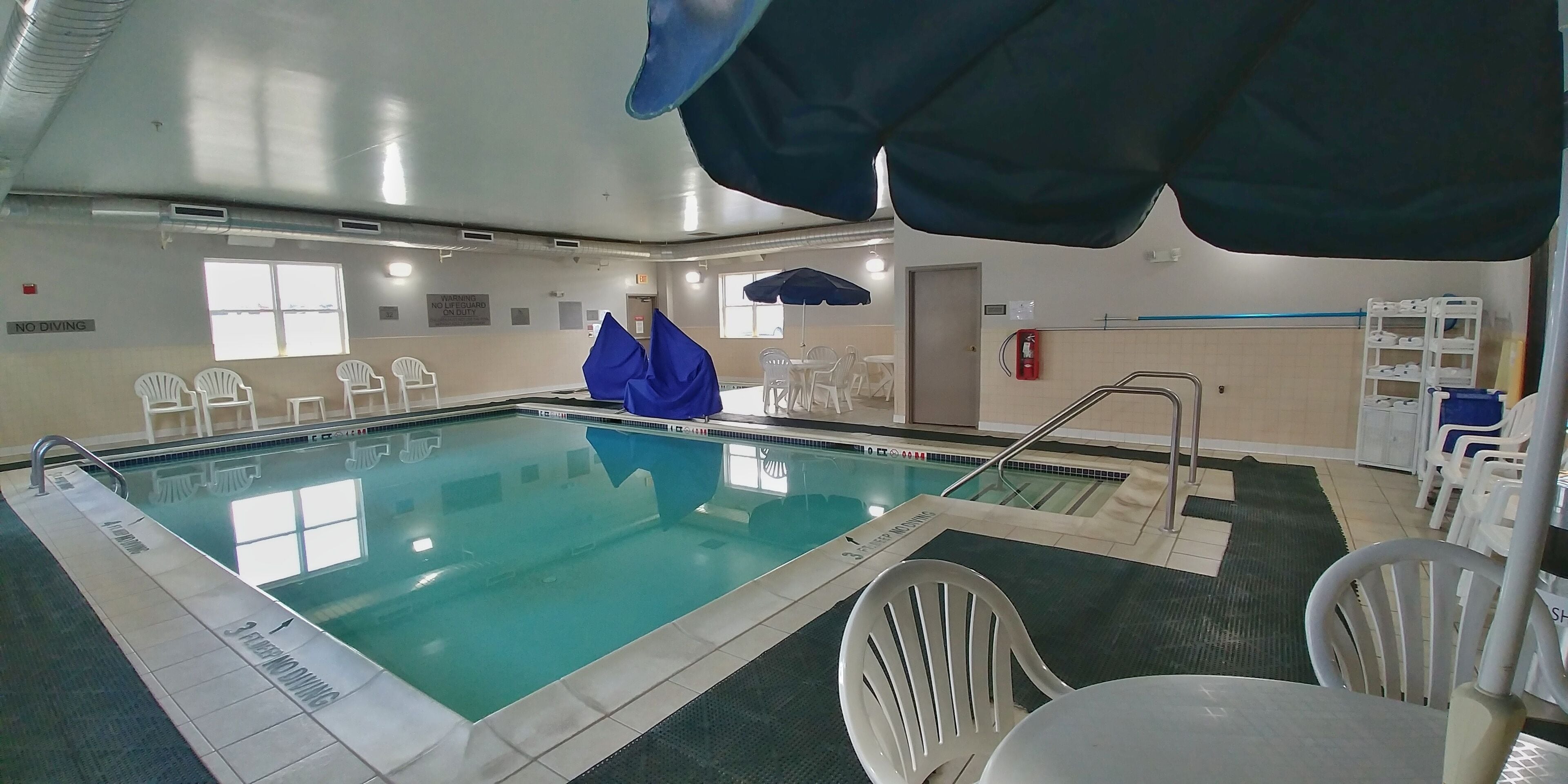 Una piscina con agua cristalina, rodeada de sillas de plástico blancas y una sombrilla azul.
