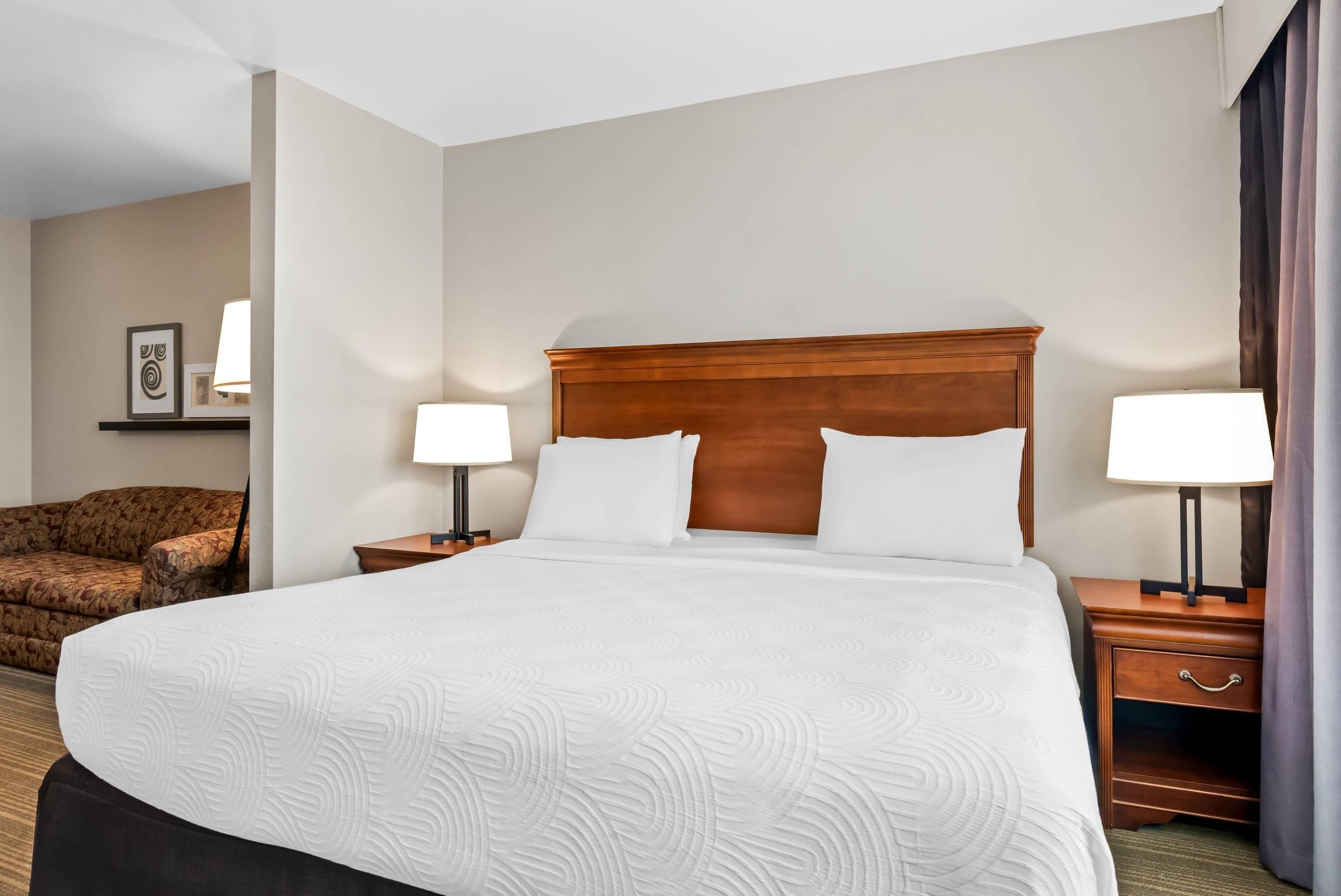 Quarto de hotel com uma cama grande, duas mesas de cabeceira com abajures, uma cabeceira de madeira, um sofá e um quadro na parede.