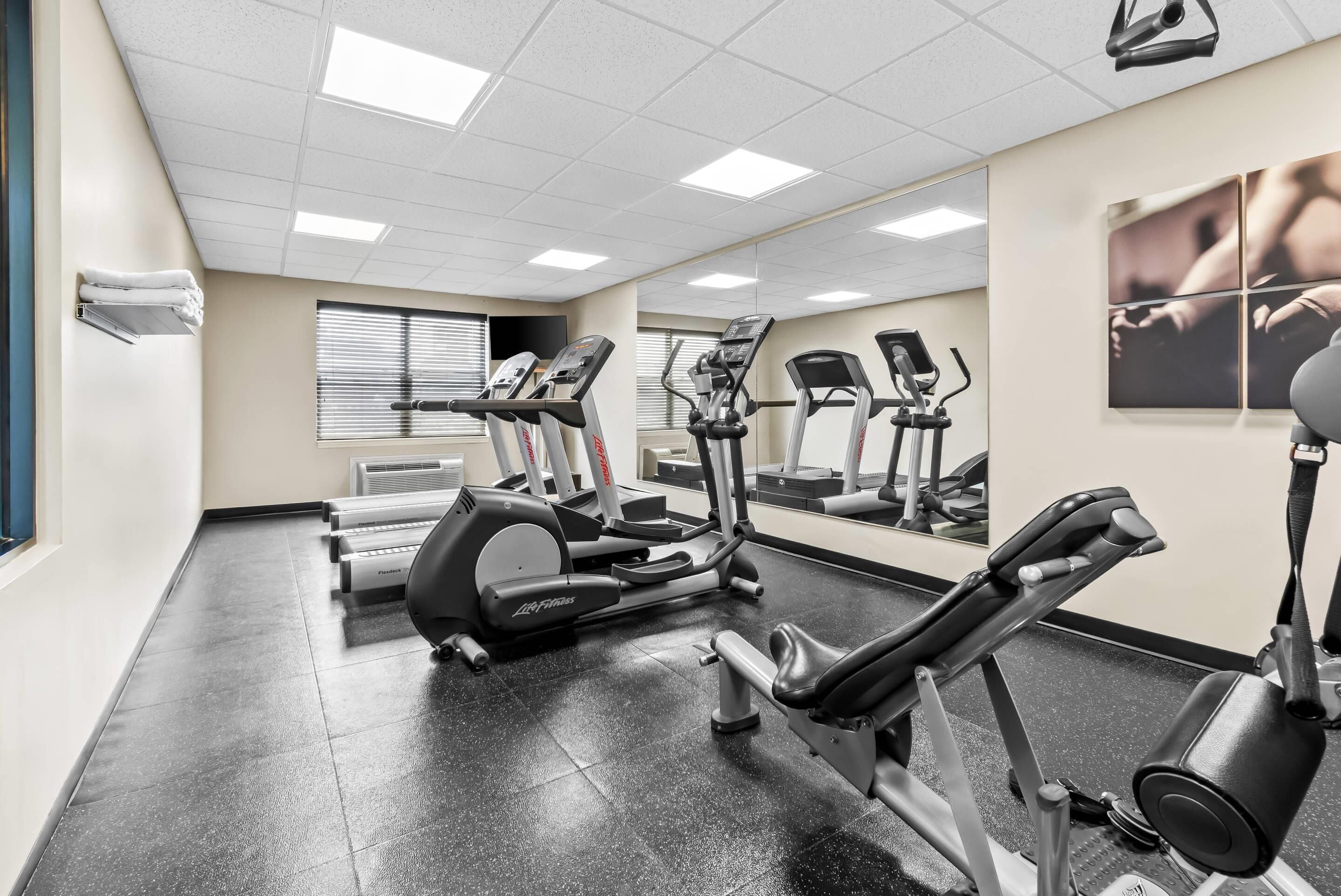 A gym with various exercise machines, a large mirror, and a window with blinds.