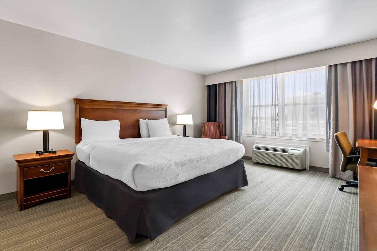 A hotel room with a bed, bedside tables, a desk, an air conditioning unit, and a large window with curtains.