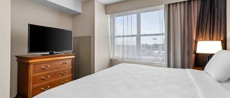 A hotel room with a large bed, a television on a wooden dresser, a window with curtains, and a lamp on a bedside table.