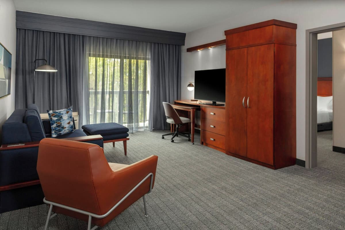 Ein Hotelzimmer mit Sofa, Sessel, Schreibtisch, Fernseher, Kleiderschrank und einem großen Fenster mit Vorhängen.
