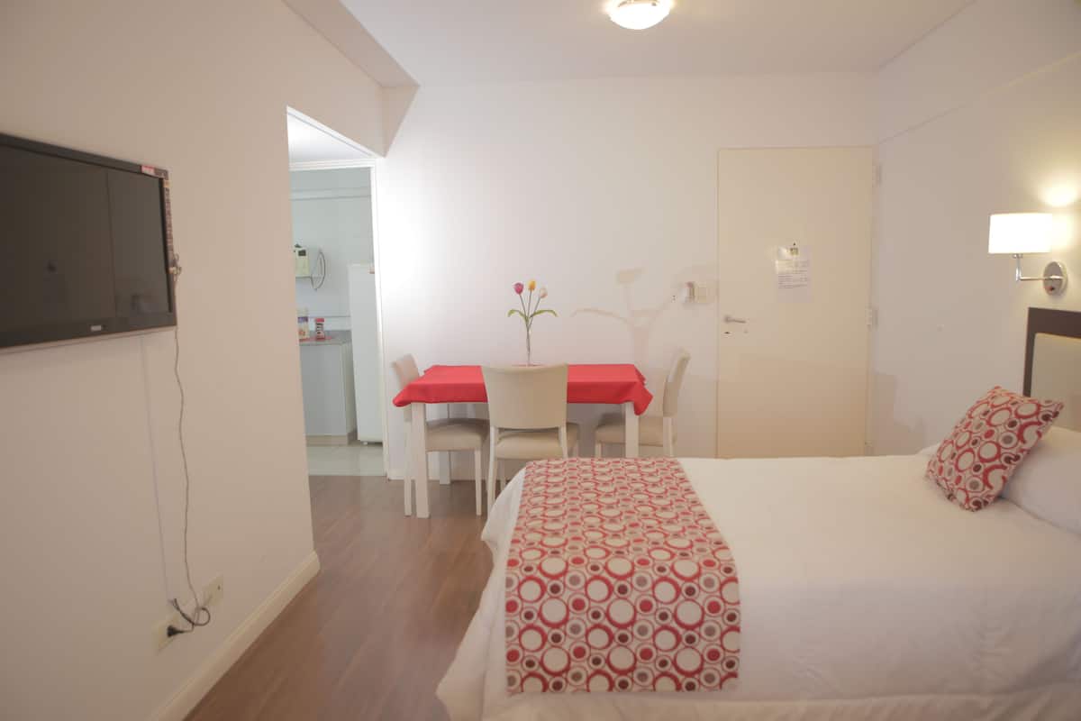 A hotel room with a bed, a desk with a red tablecloth, a chair, and a small table with a vase of flowers.