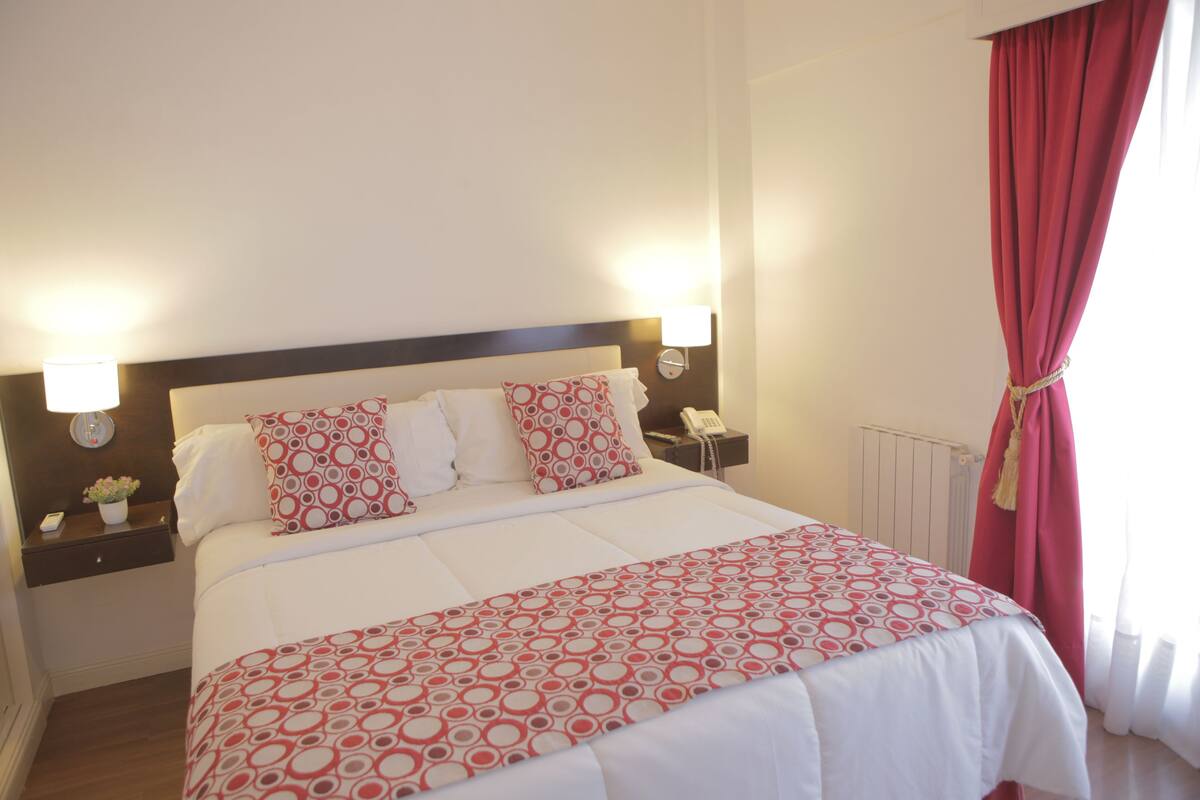 A neatly made bed with a patterned bedspread, two bedside lamps, a nightstand with a phone, and a window with red curtains.