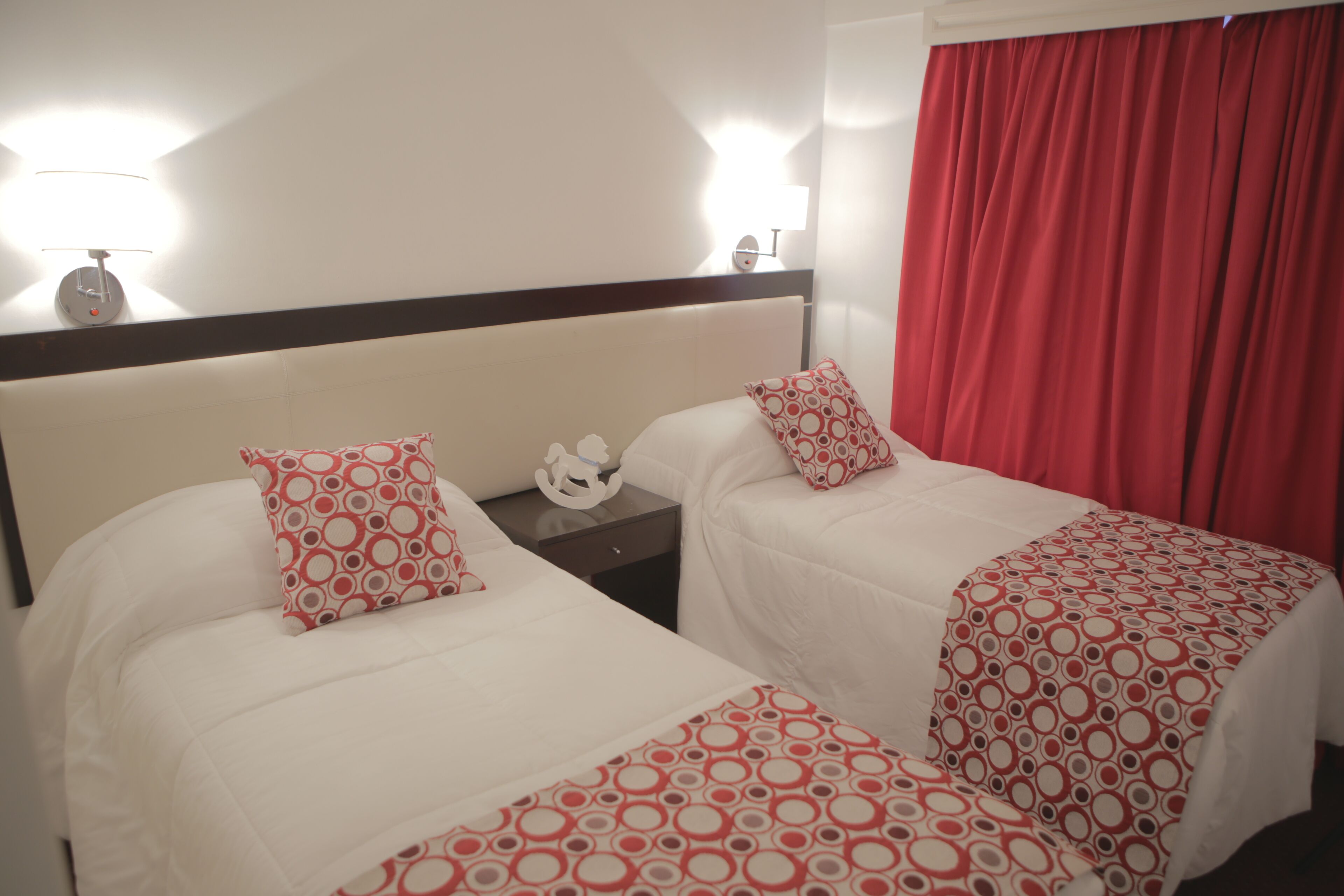 Habitación de hotel con dos camas, cada una con una colcha blanca y motivos circulares rojos. Hay dos mesitas de noche con lámparas, una cortina roja y una pared blanca con un aplique.