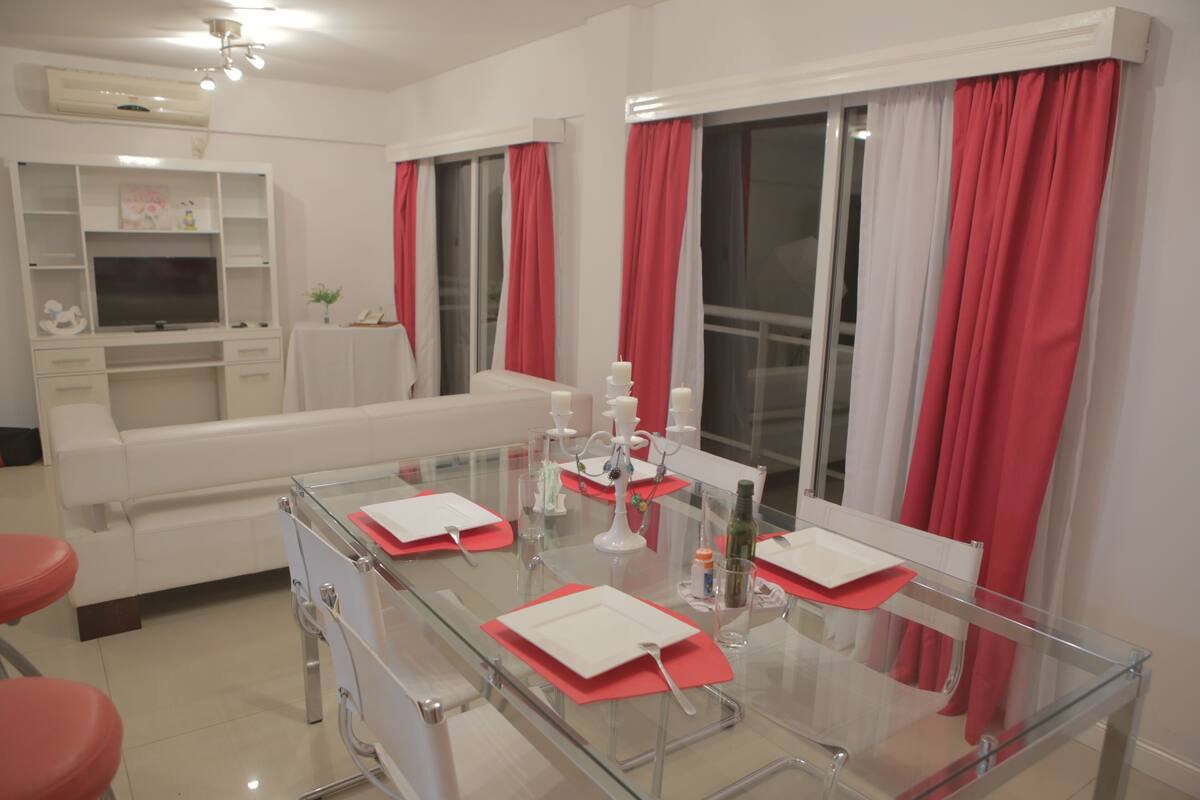A modern living room with a glass dining table, white sofa, and red curtains.
