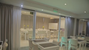 A modern hotel dining area with white tables and chairs, large windows with curtains, and a view into the dining room.