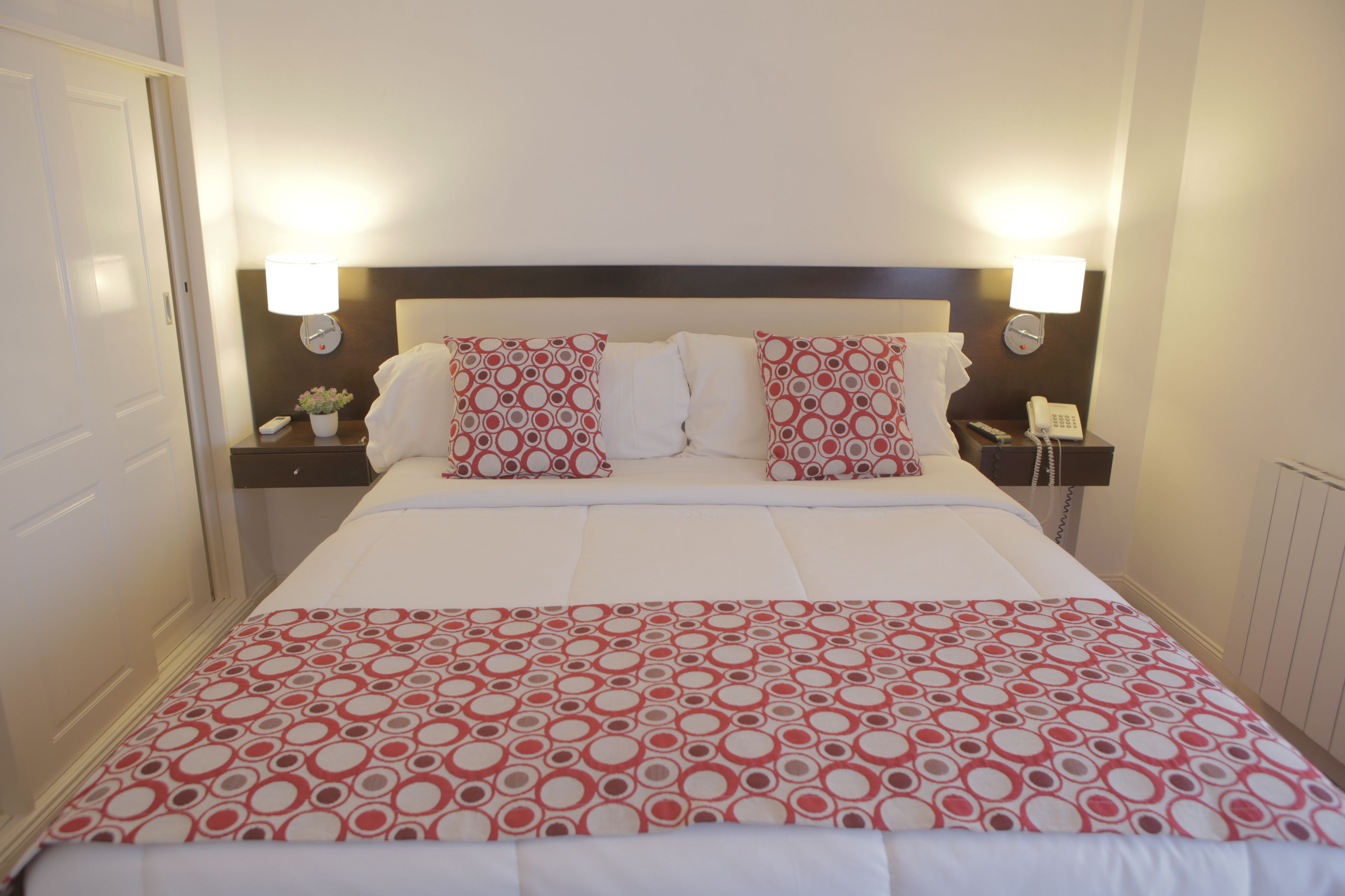 A neatly made bed with a patterned bedspread, two bedside lamps, and a nightstand with a phone and a vase of flowers.
