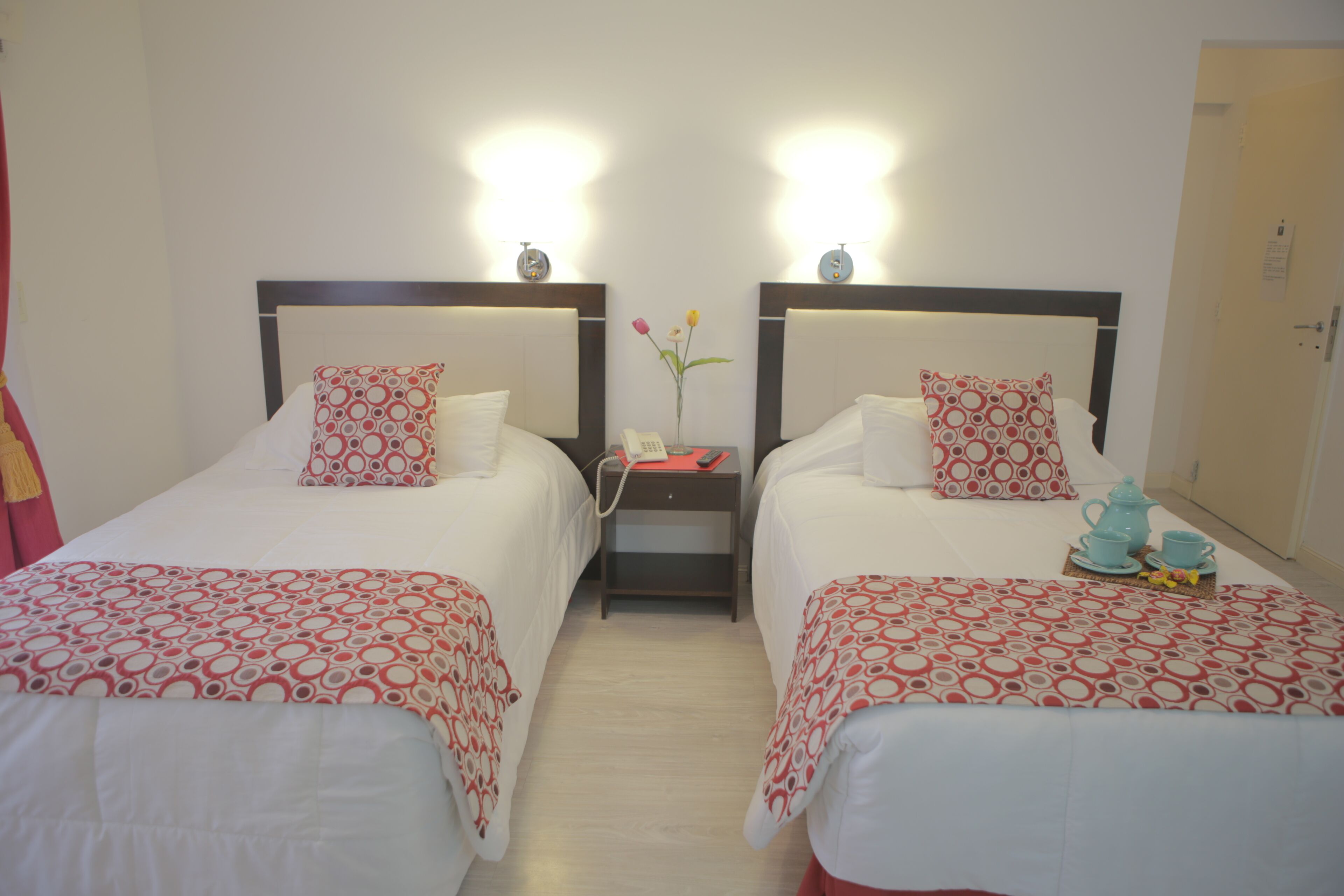 A hotel room with two beds, each with a patterned bedspread, a bedside table with a lamp, and a vase with flowers.