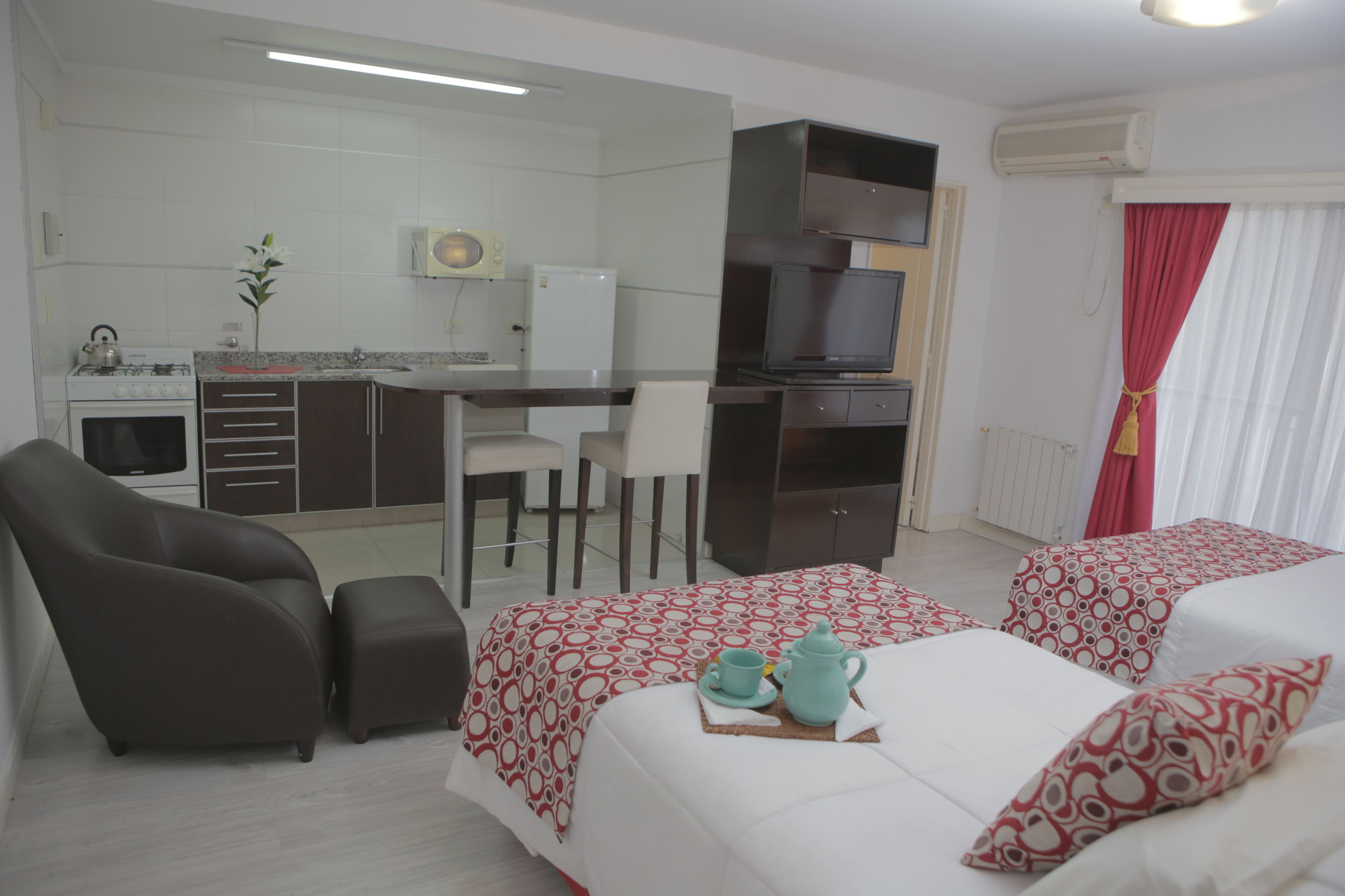 A hotel room with a kitchenette, a TV, two beds with patterned bedding, a sofa, and a dining table.