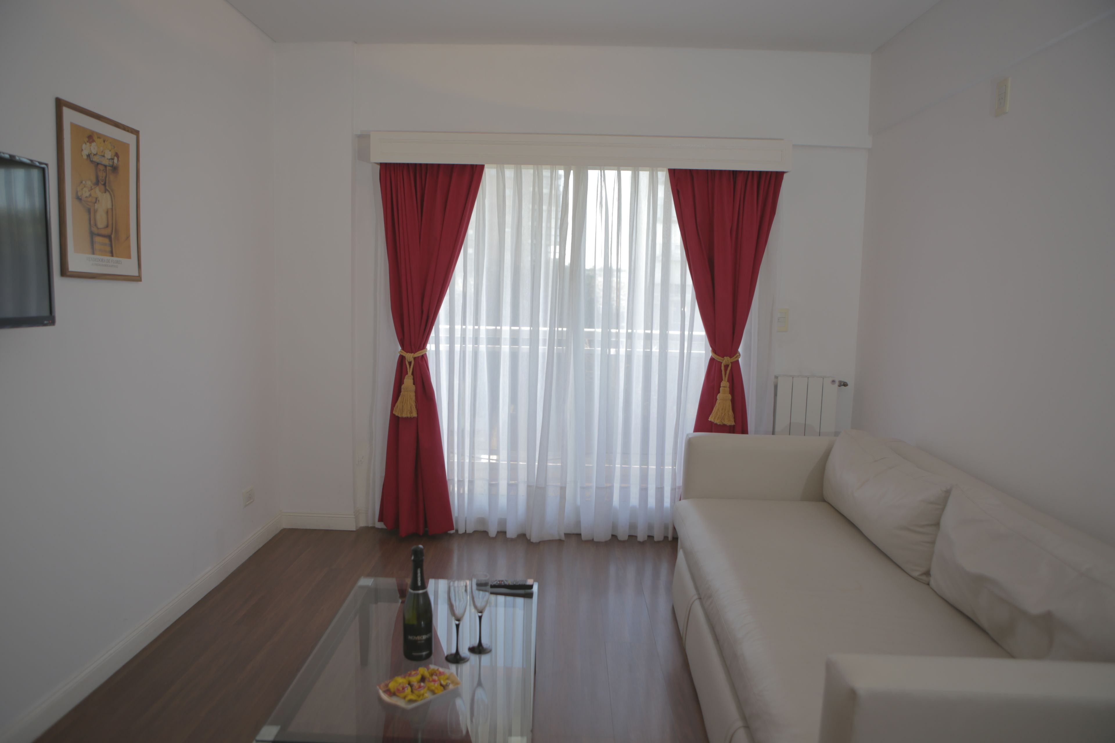A living room with a white sofa, a glass coffee table with a bottle and glasses, and red curtains.