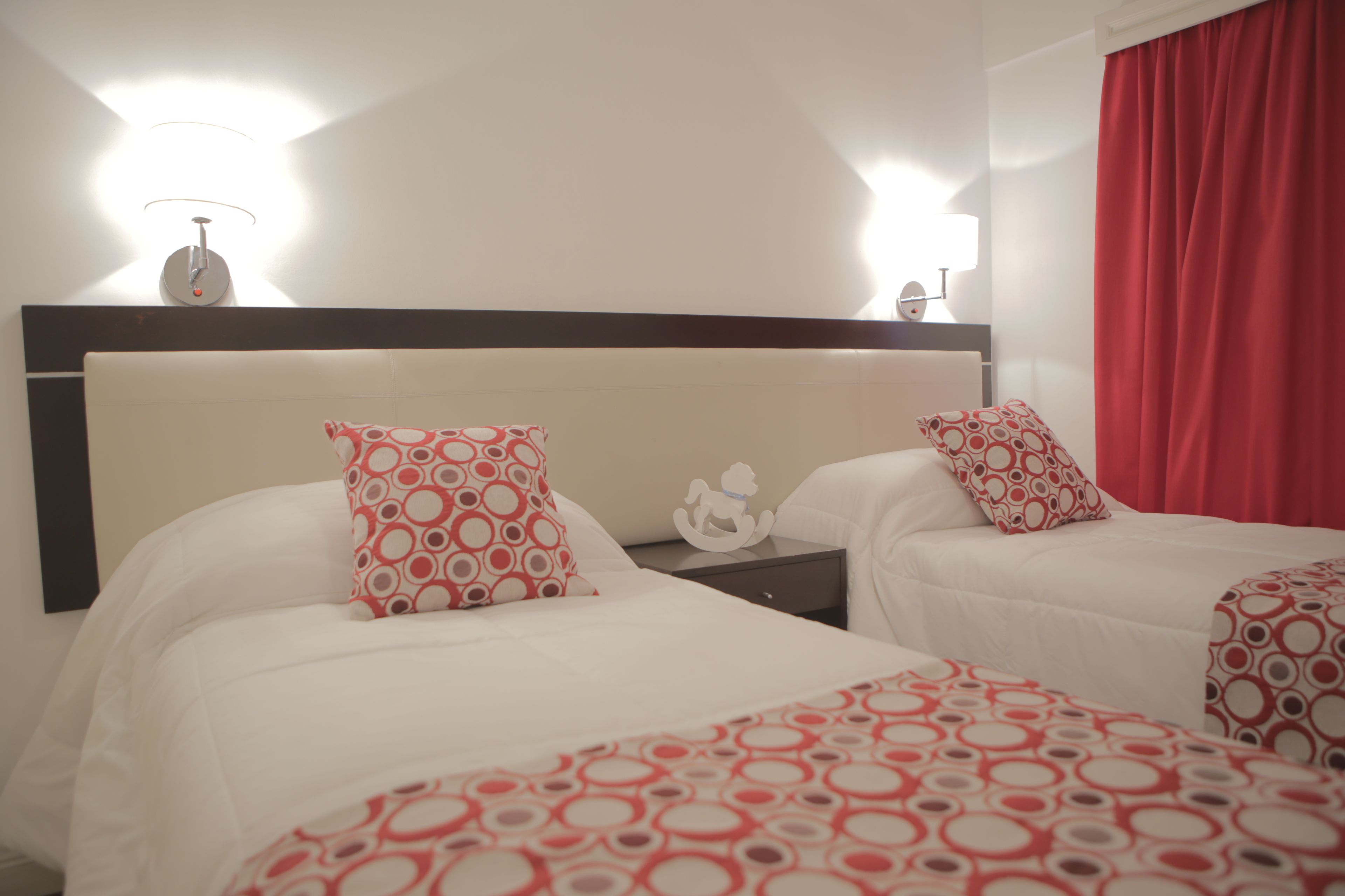 A hotel room with two beds, each with a red and white patterned bedspread, a nightstand with a lamp, and a red curtain.