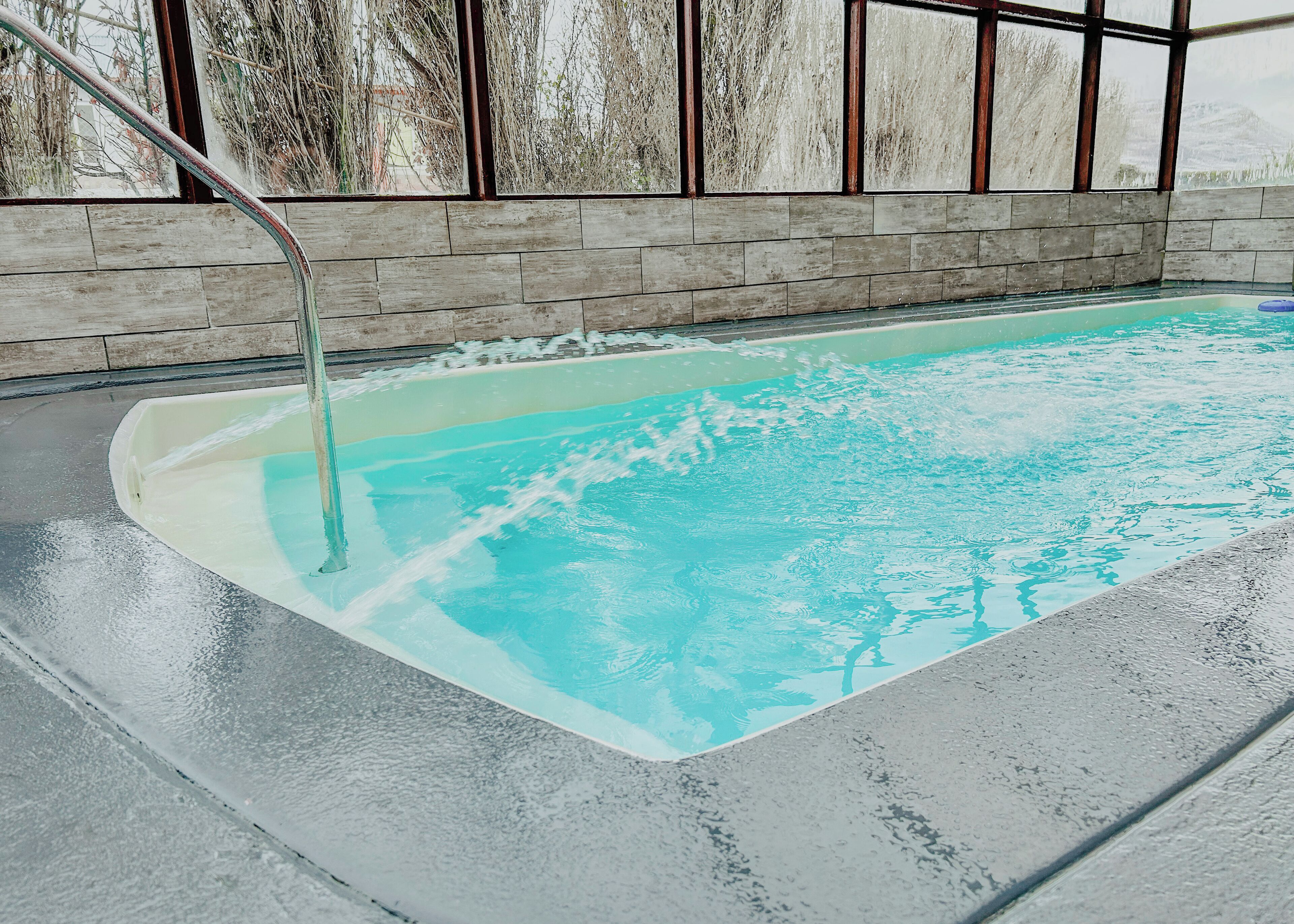 Una piscina con un elemento acuático de agua cristalina y azul, rodeada por una terraza con baldosas y una pared de madera con ventanas amplias.