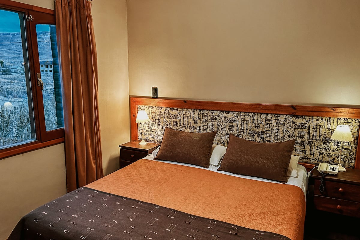 Habitación de hotel con una cama grande, una mesita de noche con lámpara, un teléfono y vistas al exterior.