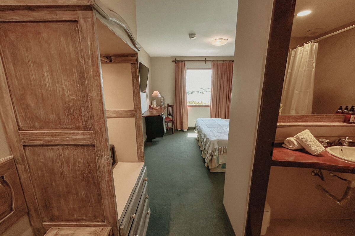A narrow hallway leading to a bedroom with a bed, a desk with a lamp, and a bathroom with a sink and mirror.