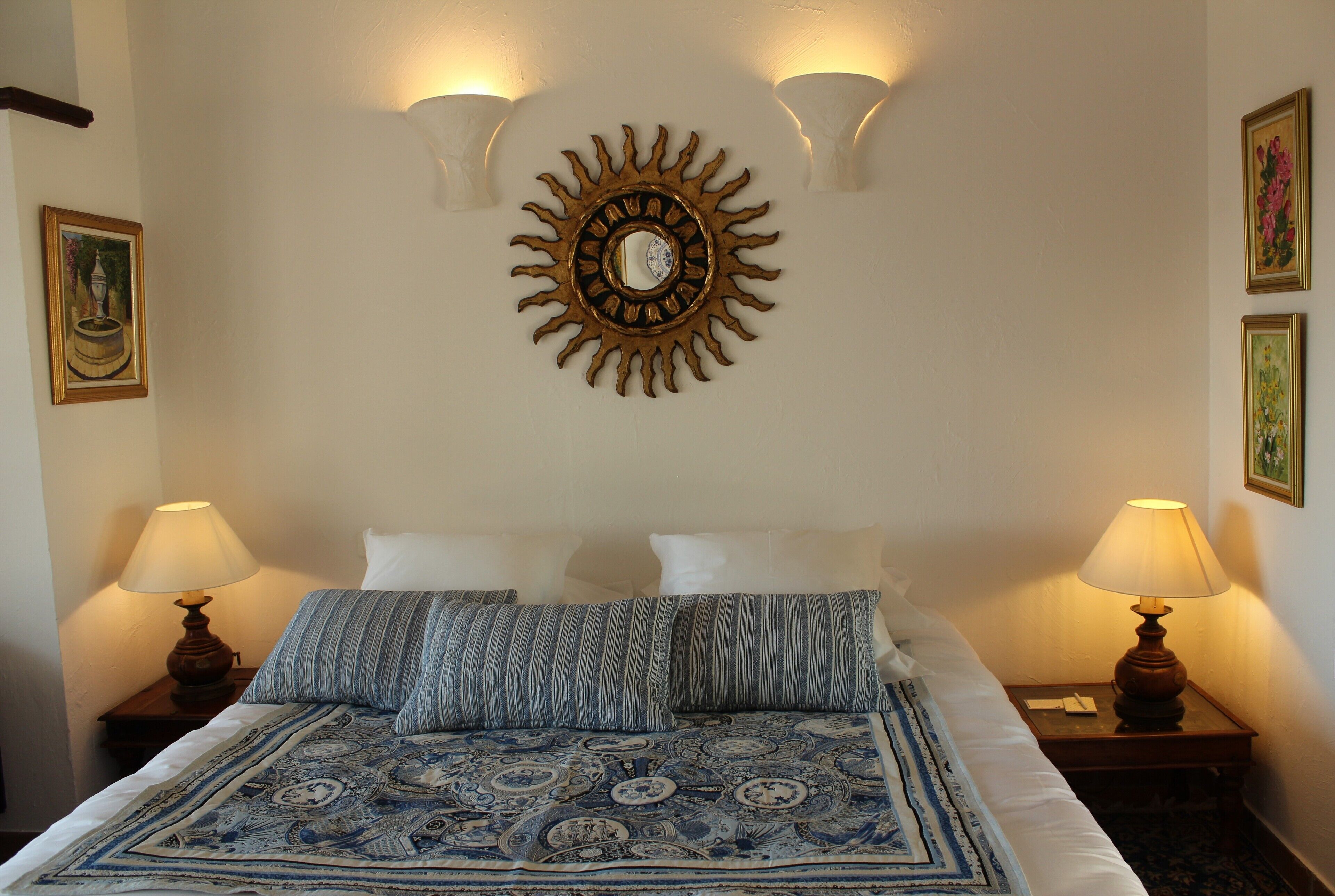 Un lit bien fait, recouvert d’une couvre-lit à motifs, deux lampes de chevet, une décoration murale représentant un soleil et des œuvres d’art encadrées accrochées aux murs.