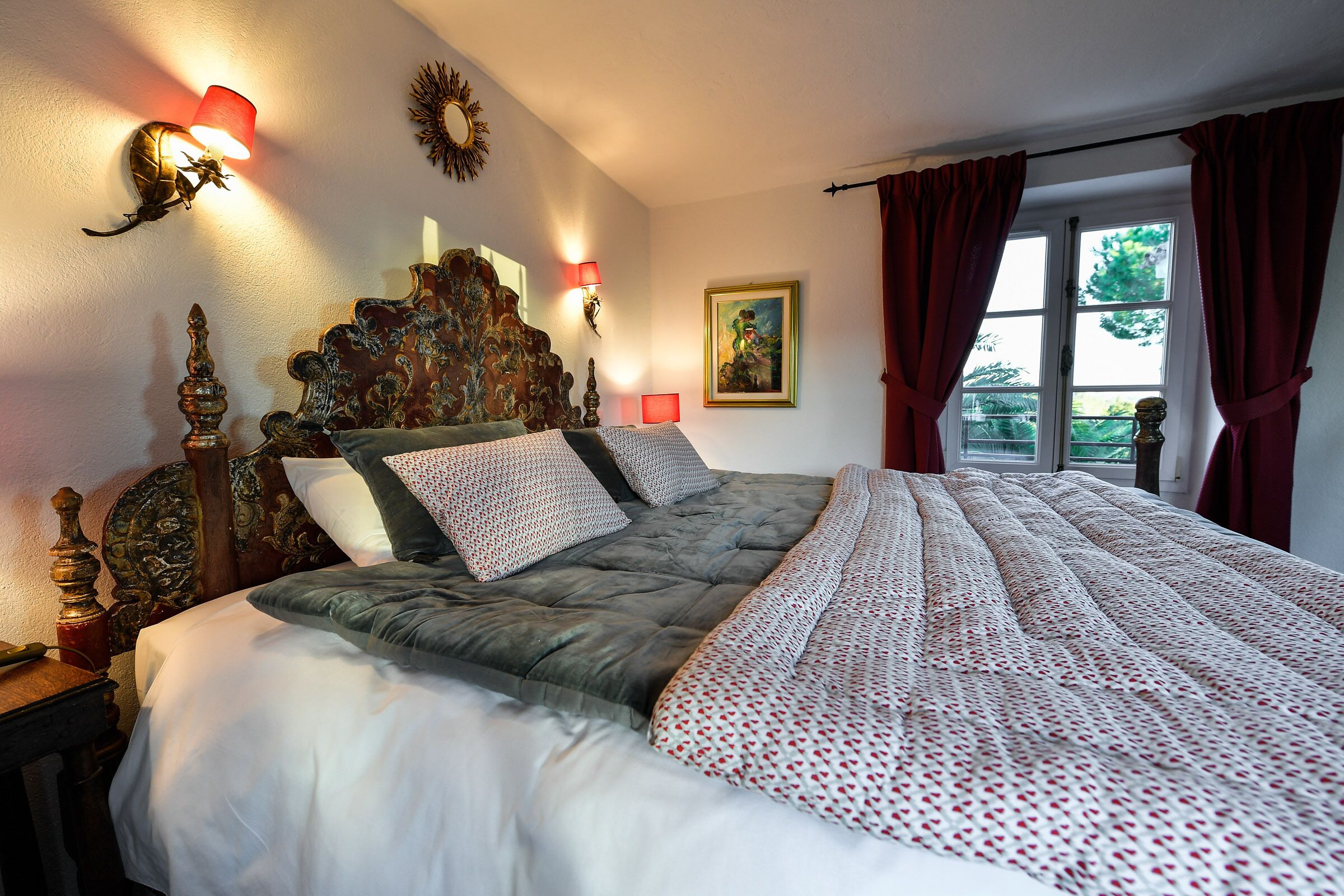 A bedroom with a large bed, ornate headboard, wall sconces, a framed painting, and a window with curtains.
