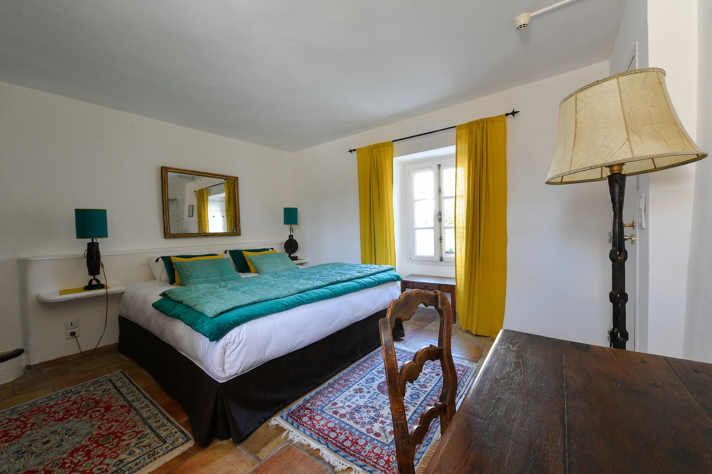 A bedroom with a bed, bedside lamps, a wooden chair, a table, a window with yellow curtains, and a lamp on a stand.
