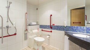 Une salle de bain avec des toilettes blanches, un lavabo et une douche. Des barres d’appui rouges sont installées près des toilettes et du lavabo.