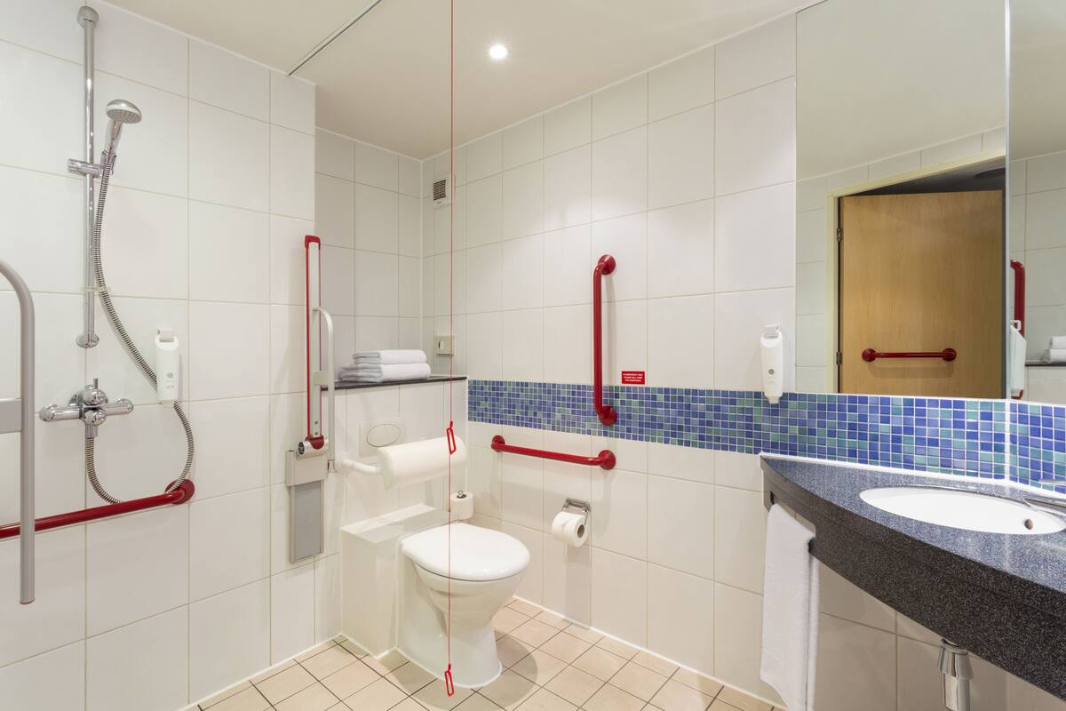 A bathroom with a white toilet, a sink, and a shower. There are red grab bars installed near the toilet and sink.
