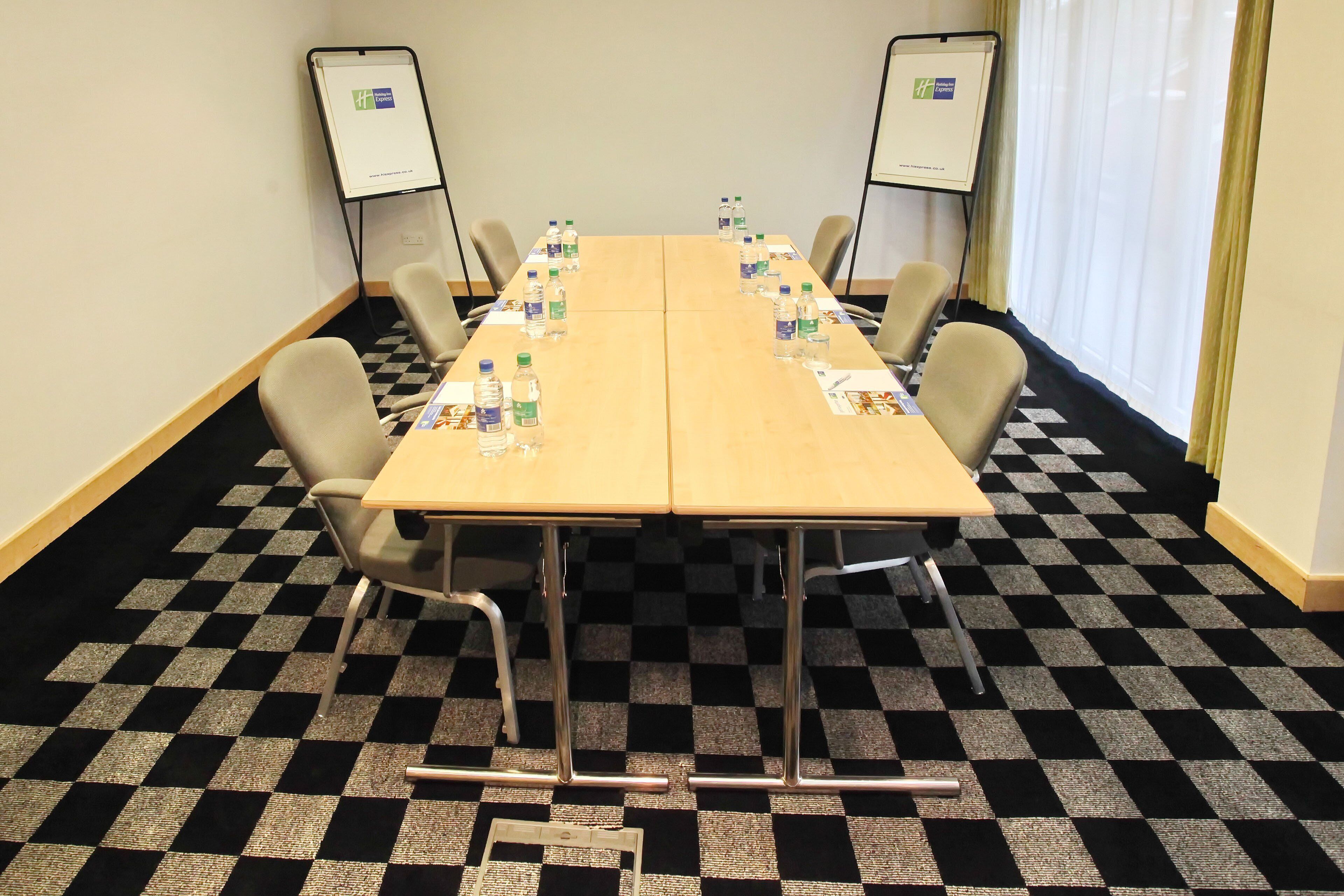 A conference room with a checkered floor, a long table with chairs, water bottles, and two flip charts on stands.