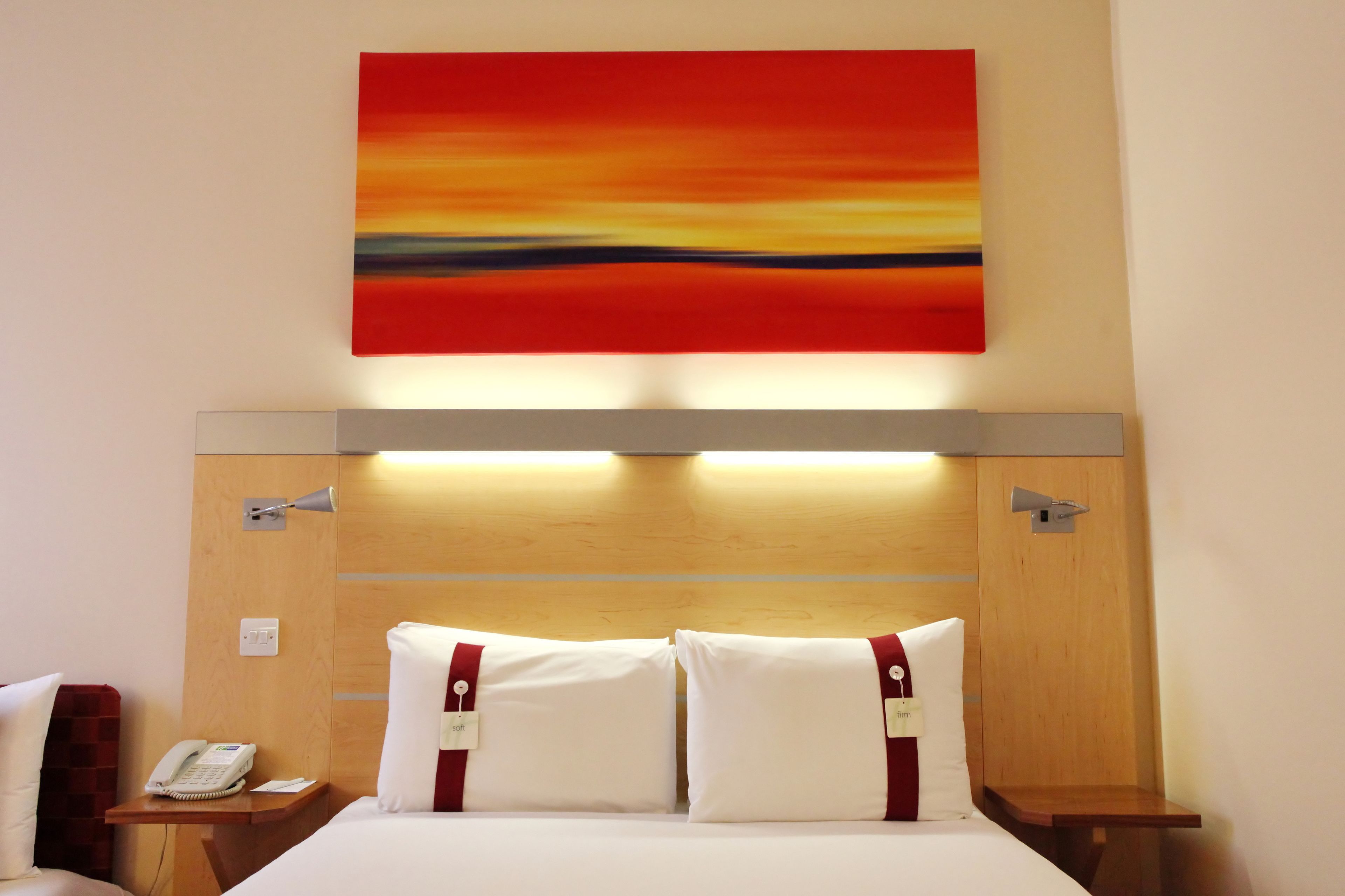 A hotel room with a wooden headboard, a bed with white bedding and red accents, a bedside phone, and a wall-mounted artwork.