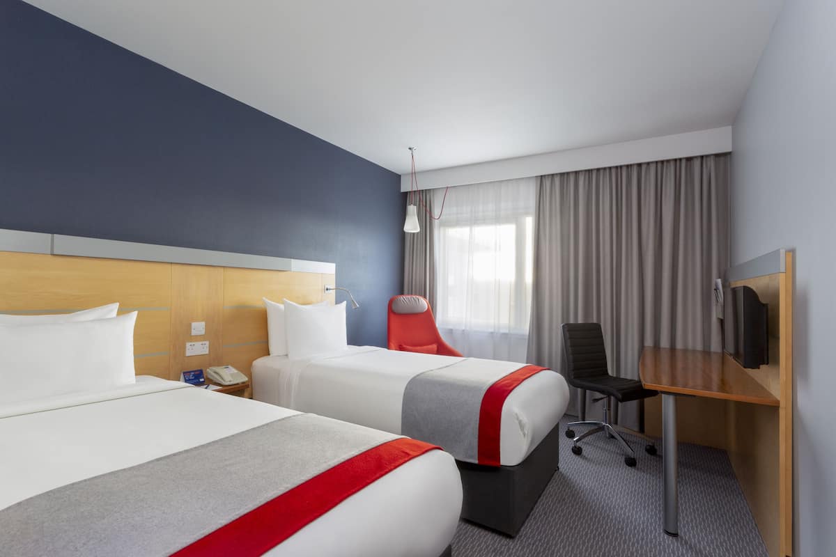 A hotel room with two beds, a desk, a chair, a TV, and a window with curtains.