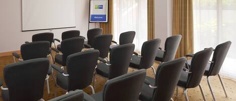 Un salón de conferencias con filas de sillas, una pantalla de proyector y una pantalla mostrando 'Bienvenido'.