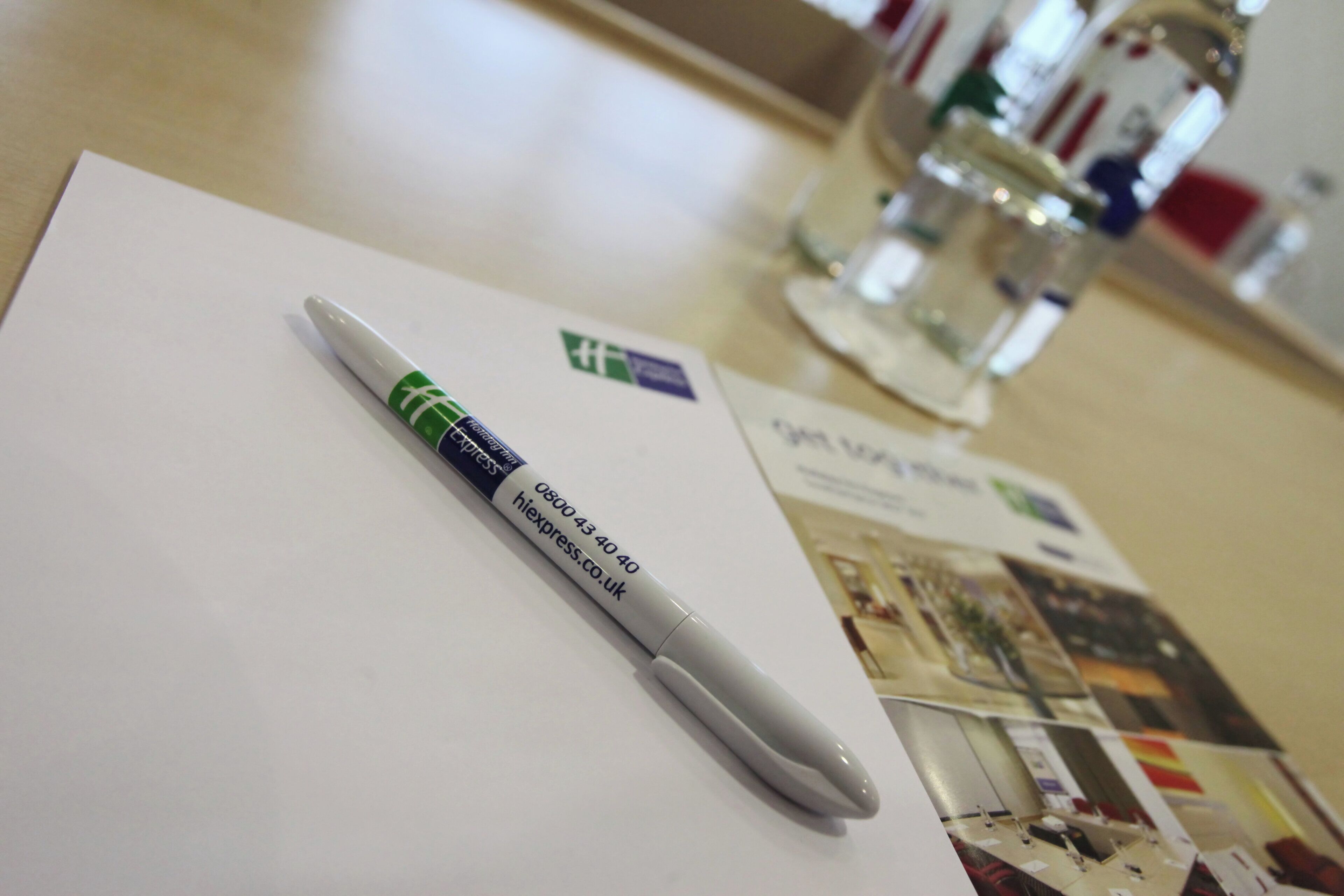 A white notepad with a pen on top, a glass of water, and a blurred background with a poster.
