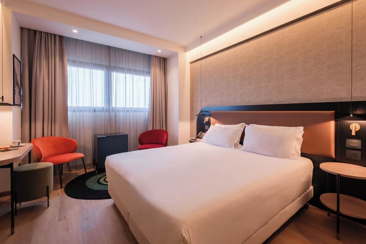 A hotel room with a large bed, a desk with a chair, a small table, and a red chair.