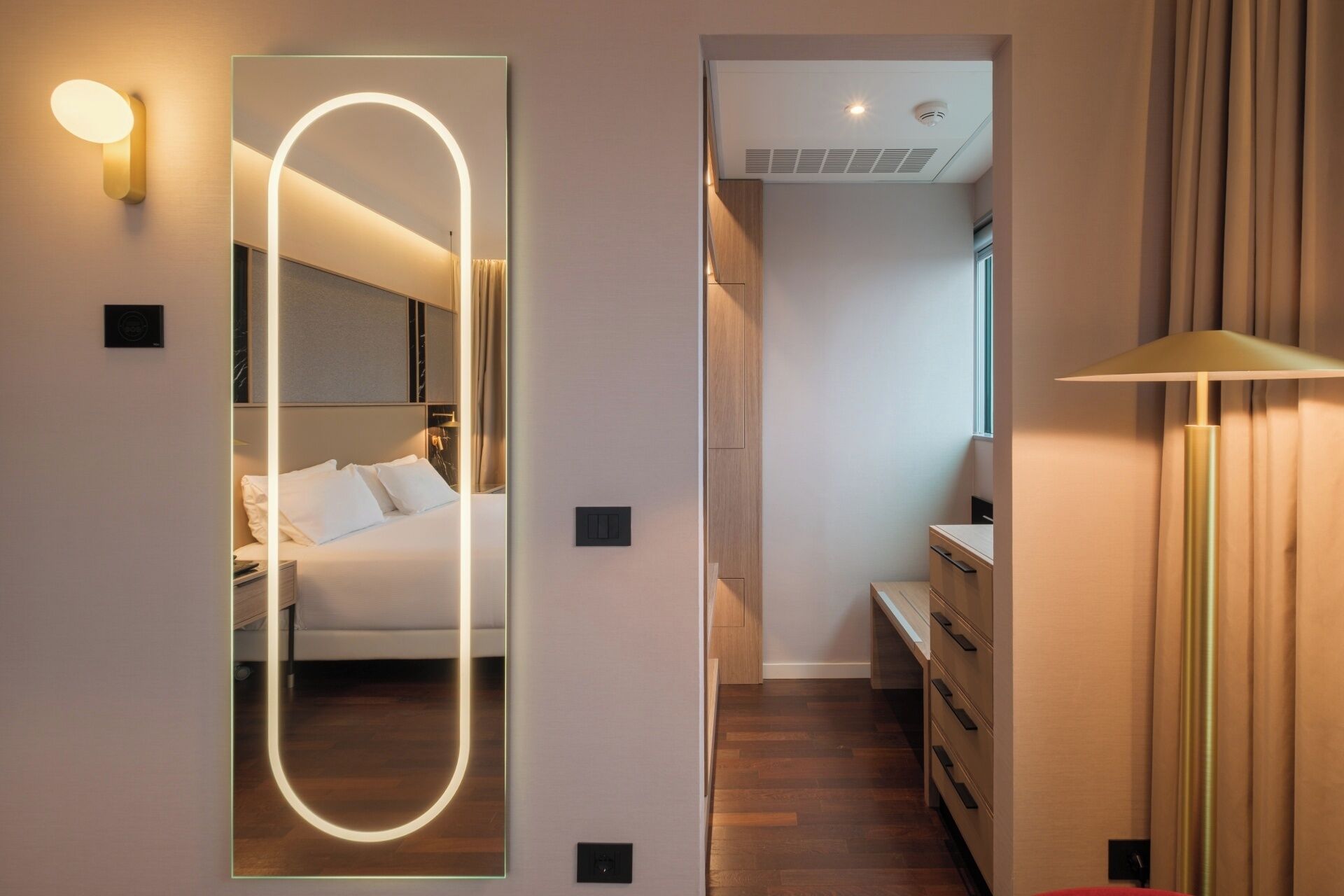 A modern hotel room with a large mirror, a bedside table, a lamp, and a wooden floor.