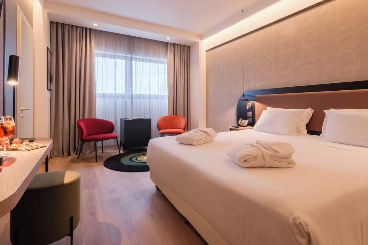 A hotel room with a large bed, two chairs, a desk with a lamp, and a view of the outdoors through the window.